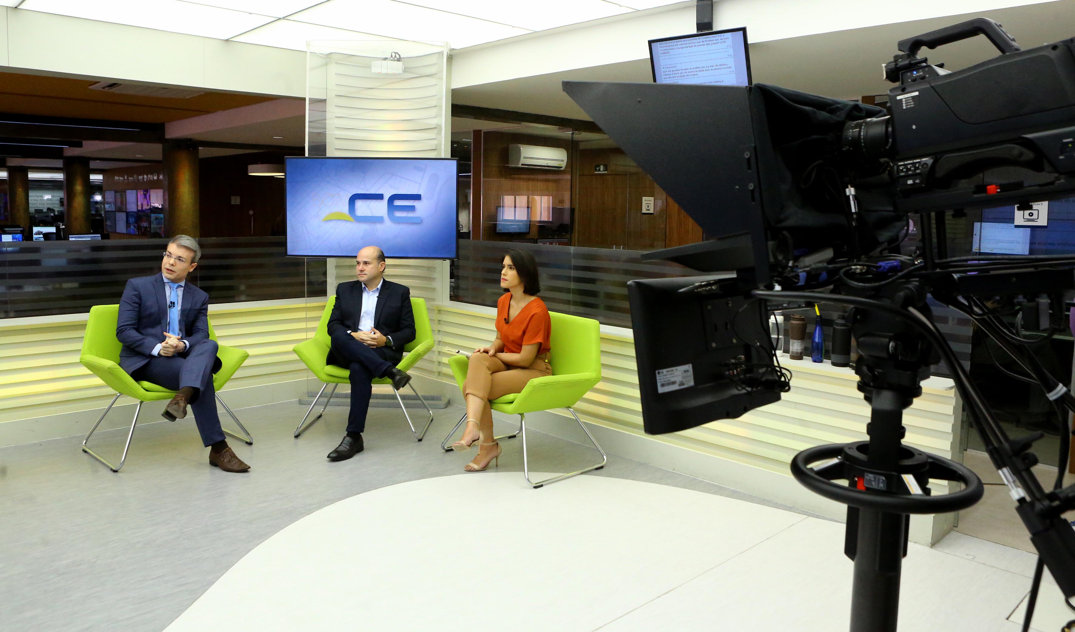 prefeito sentado entre dois apresentadores em estúdio de televisão com câmera ao lado